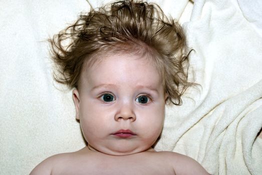 Cute little kid with shaggy hair, lying on the bed looking at the camera.