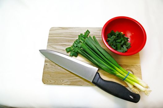 A cutting Board with a knife, bowl and a bunch of green onions