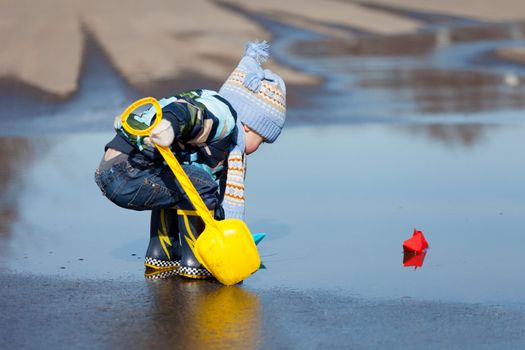 Little boy plays with paper ships in a spring puddle