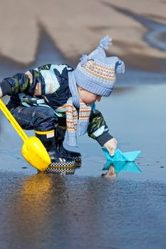 Little boy plays with paper ships in a spring puddle