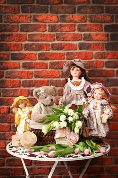 Some porcelain dolls and teddy bears on a table against a wall from a red brick