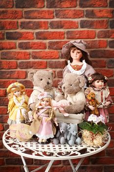 Some porcelain dolls and teddy bears on a table against a wall from a red brick