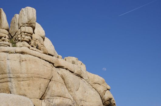 Iconic grey rock formation only found at Joshua Tree National Park, California - against a blue sky with moon and aeroplane 'chem' trail visible in the sky. Copy  space.