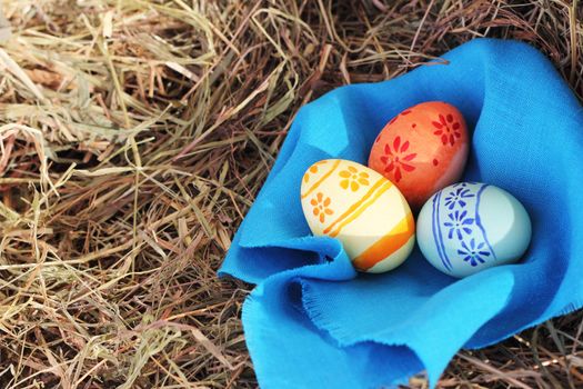Three colored Easter eggs and blue textile on hay at sunny day