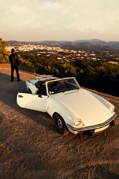 View of a young male with a jacket next to his white convertible car.