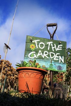 A portrait format image set at a low angle, of a terracotta colored flower pot with a garden fork and a hand painted wooden sign with garden themed images. Set in a small city garden against a bright blue sky background.