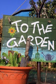 Close up of a hand painted wooden sign with garden themed illustrations and text, arranged with a terracotta colored flower pot to foreground with a small garden trowel.