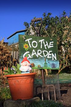 A portrait format image of a terracotta colored flower pot and garden gnome in front of a garden fork and hand painted wooden sign. Set in a small city garden against a bright blue sky background.