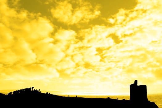 a silhouette of a crowd on a hill beside old castle with a view of the sea and sunset