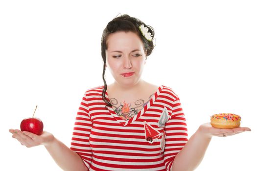 A young, woman, dressed in Rockabilly style, trying to decide whether to eat the tempting, sweet donut, or the healthy, fresh apple.