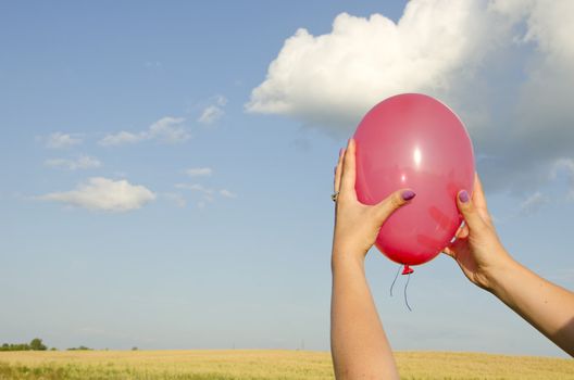 Woman hand holding red balloon on background of sky and farm fields.