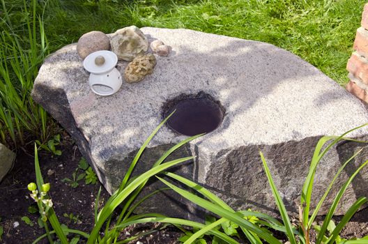 Decorative ancient stone with water in hole and objects on it in garden.