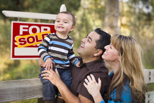 Happy Mixed Race Couple with Baby in Front of Sold Real Estate Sign.