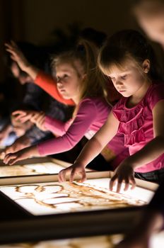 Girl paint with sand on table by her fingers in classroom with friends