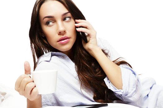 beautiful woman in bed holding coffee while calling with her phone on white background