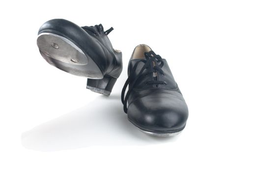 A pair of used black tap shoes with laces, one on the ground and one raised up, ready to step.  White background, shadows visible.