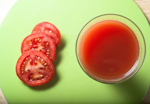 Overhead shot of tomato juice in a long, tall glass beside three slices on a fresh green circular mat, resting on  birch wood table.  