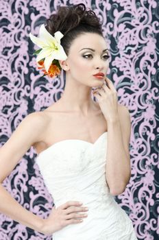 Young adult bride in white wedding dress posing over magenta wall. Her hair decorated with lily flowers