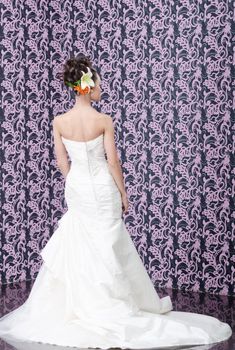 Young adult bride in white wedding dress posing over magenta wall. Her hair decorated with lily flowers