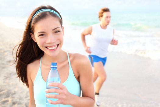 Runner woman drinking on beach with man running in background. Happy smiling mixed race Asian / Caucasian female fitness sport model during outdoor workout.