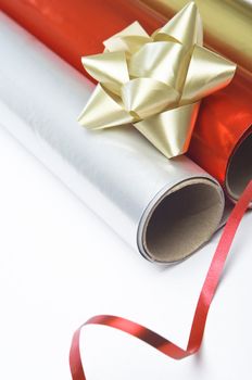 Close up, angled shot of gift wrapping paper rolls, rosette and ribbon on a white surface.