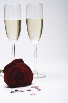 Two champagne glasses (flutes) have focus, with dark red rose and a scattering of heart confetti in soft-focus in the foreground.  