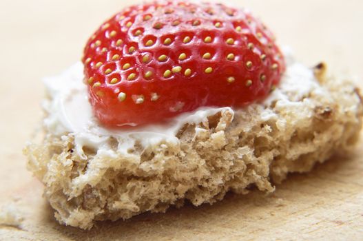 A heart shaped piece of bread with topping of cream cheese and half a strawberry, on a wooden choppinng board.