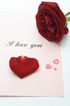 An opened Valentine's card with a red rose, a single petal and heart-shaped confetti, on an off-white surface.  