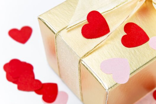 Close up (macro) of a gold gift box, tied and bowed with organza ribbon and scattered with pink and red paper heart confetti.  Background is off-white.