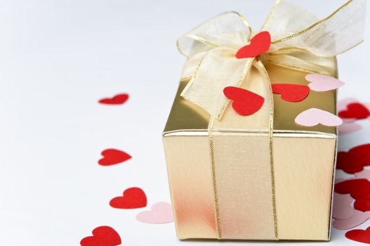 Close up of a gold gift box, tied and bowed with organza ribbon and scattered with pink and red paper heart confetti.  Background is off-white.