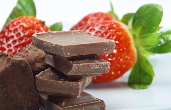 Close up of a stack of chocolate pieces with dusted truffles to the left, and strawberries in soft focus in the background.  The surface is a grey-blue china plate.