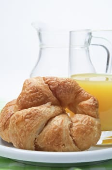 A continental breakfast setting.  A croissant on a white china plate with a green gingham tablecloth beneath in the foreground, with glass and jug of orange juice in the background.  