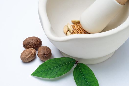 Close up of a stoneware pestle and mortar containing a mixture of whole spices, with nutmegs and bay leaves on the surface outside it. Horizontal (landscape) orientation.