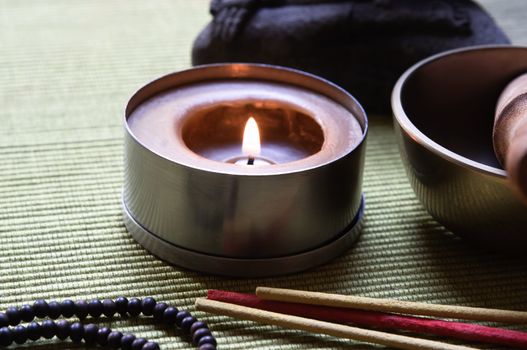 Close up of a collection of Buddhist's objects (candle, josticks, prayer beads and singing bowl), with part of a Buddha statue in the background. Placed on an olive green ribbed mat.