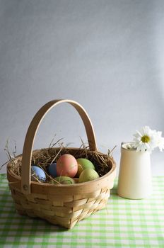 Wooden basket of decorated Easter eggs and straw on a gingham table cloth with a jug of flowers to the side.   Portrait (vertical) orientation.