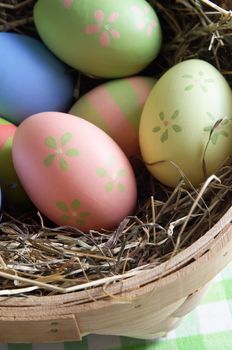 Close up of colourful, hand painted Easter eggs nestling in straw inside a basket on a gingham table cloth. Shot from above.