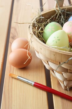 Hand painted Easter eggs in a basket filled with straw with natural, unpainted eggs and paintbrush resting on a wood plank table top. Vertical orientation.