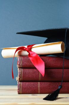 A stack of old, worn books with a mortarboard and ribbon tied scroll on top, placed on a wooden table with a grey background.