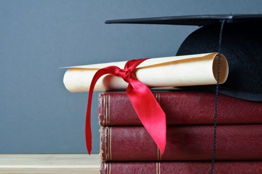Close up of a mortarboard and graduation scroll on top of a pile of old, worn books, placed on a light wood table with a grey background.