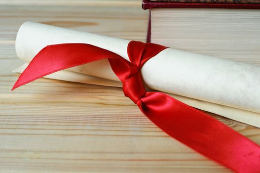 A scroll tied with red ribbon on a wooden surface in front of a book, to illustrate diploma or degree.
