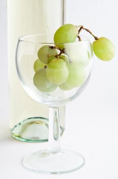A wine glass filled with white (green) grapes, standing in front of a cropped bottle of white wine.