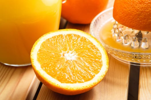 An orange half in the foreground with squeezer, whole orange and jug of juice behind on a wood planked table.