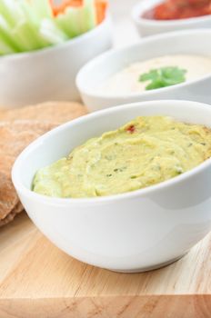 Guacamole, hummus and salsa dips in white bowls on a wooden board with wholemeal pitta bread and crudites cropped at left frame.