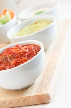 Close up of a bowl of tomato salsa, with guacamole, hummus dips and crudites in the background.