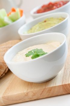 Hummus, guacamole and salsa dips in whie bowls on wooden chopping board.  Wholemeal pittas and crudites cropped at left frame.