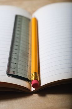 An opened student's exercise book on a wooden table, blank pages revealed.  A pencil and ruler lie vertically in the centre seam.  Vertical (portrait) orientation.