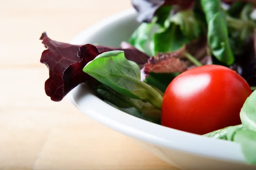 Close-up (macro) of red lettuce, rocket and cherry tomato salad in a white china bowl on a light wooden table.  Landscape (horizontal) orientation.