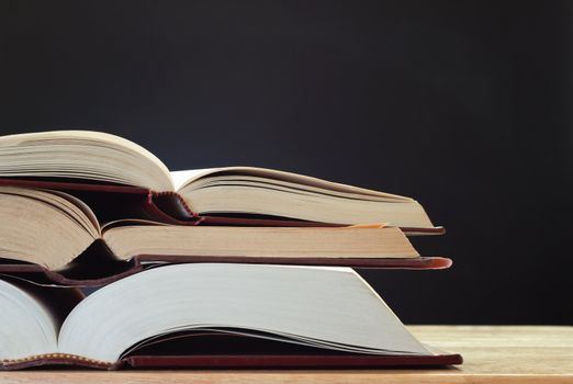 A pile of three opened books on a light wood desk, with a chalkboard (blackboard) in the background for copy space.  Landscape (horizontal) orientation.