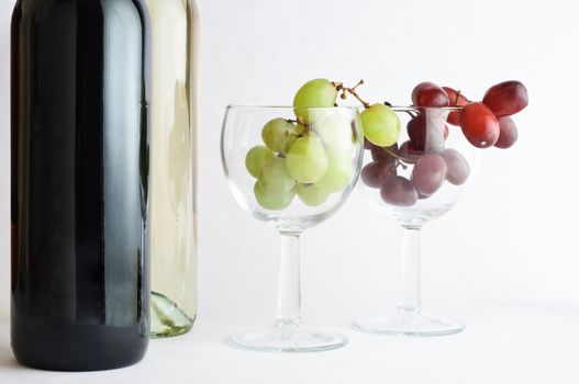 Two bottles of wine on left side of two wine glasses containing red and white grapes.