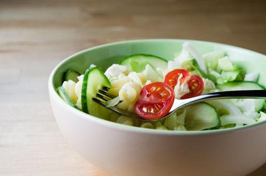 Fusili pasta with salad and fork in a bowl on a light wooden table.   